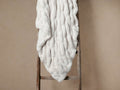 Light beige faux fur pleated throw blanket draped over a rustic wooden ladder against a warm beige wall.