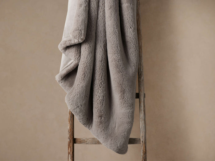 Grey faux fur oversized rabbit throw, plush and soft, draped over a rustic wooden ladder against a warm neutral wall.