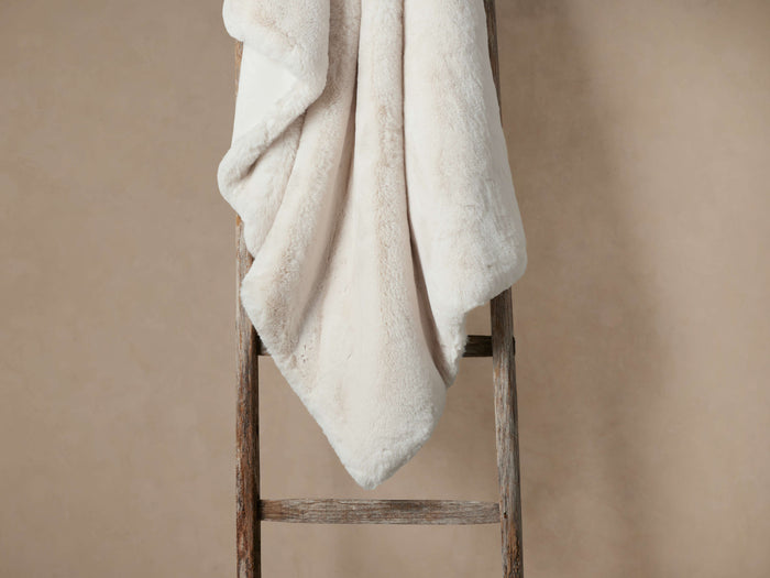 Soft cream faux fur rabbit throw draped elegantly over a rustic wooden ladder against a textured beige wall.