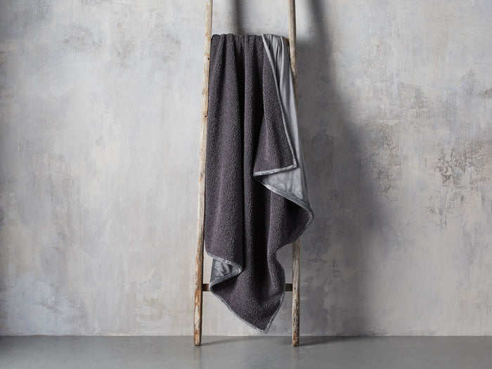 Faux Fur Sherpa Throw in dark gray, draped over a wooden ladder against a textured gray wall, showcasing its soft texture and casual elegance.
