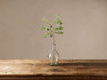 Faux olive stem with green leaves & black olives in a clear glass vase on a rustic wooden table against a beige wall.