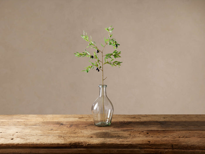 Faux olive stem with green leaves & black olives in a clear glass vase on a rustic wooden table against a beige wall.