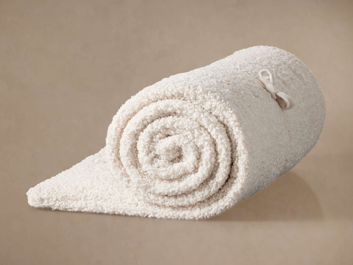 Cream Faux Sherpa Throwbed rolled up diagonally, displaying its plush, textured surface and a tie closure on a warm tan background.