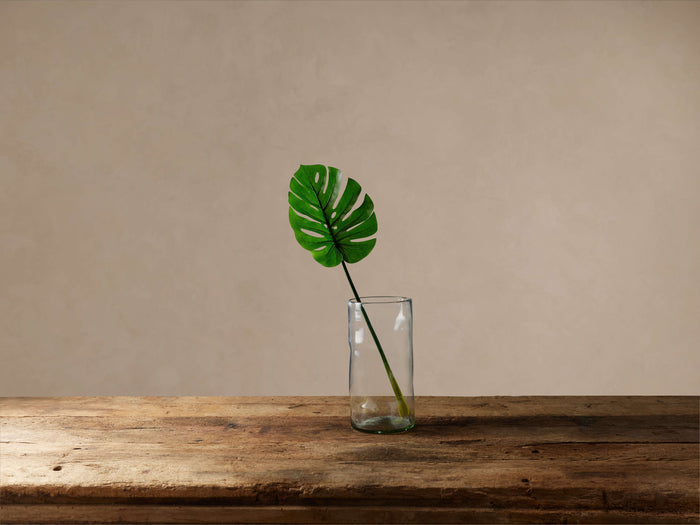 Vibrant green Faux Split Leaf Philo Stem in clear glass vase on a rustic wooden surface against a neutral background.