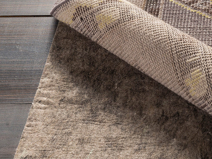 Felted Rug Pad under a patterned rug, showing its grey felt texture and no-slip rubber backing. The rug corner is lifted.