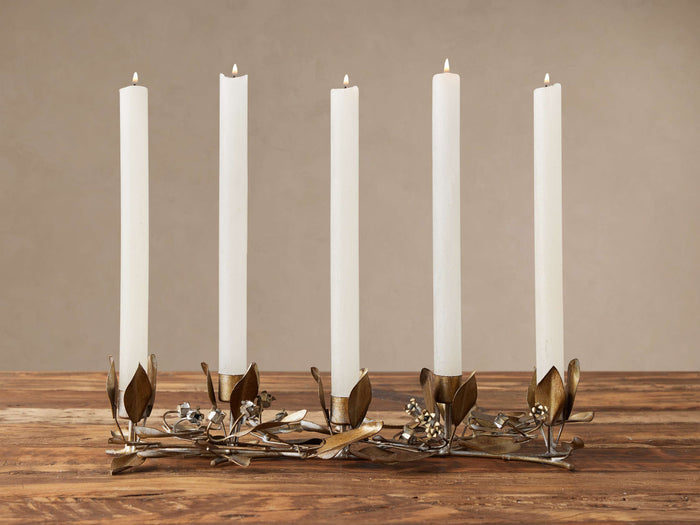 Five lit white taper candles in a long, intricately crafted champagne gold floral taper holder with leaf designs on a wooden table.
