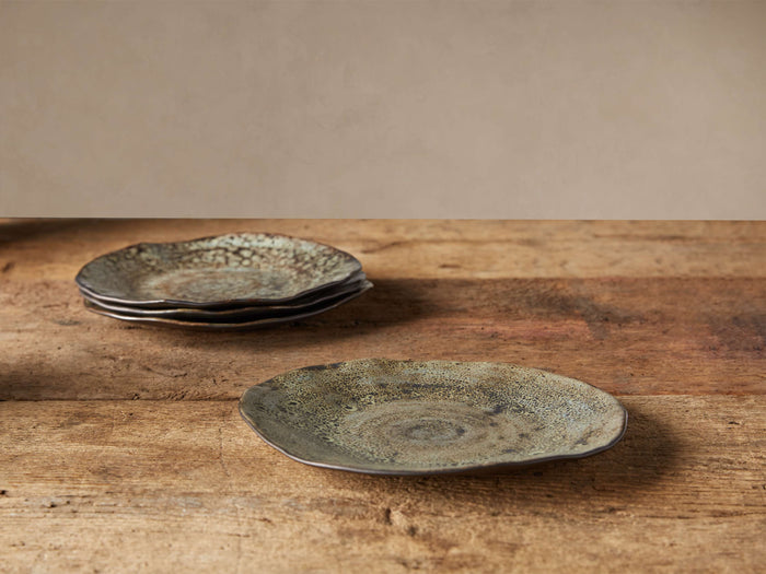 Four rustic Gallatin Dinner Plates with organic edges on a wooden table. One in front, three stacked behind, showing reactive green and brown glazes.