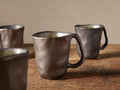 Four rustic Gallatin stoneware mugs with dark bronze reactive glaze, showing mottled greenish interiors, on wood.