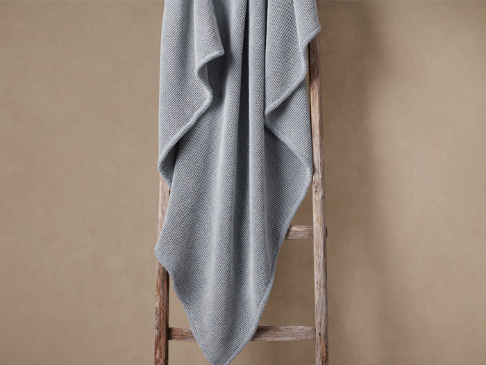 Sky blue garment-dyed throw blanket with waffle texture draped casually over a rustic wooden ladder against a light brown wall.