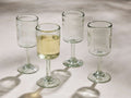 Four Granada recycled glass wine glasses, cylindrical and stemmed. One holds yellow wine, all cast shadows on a light surface.