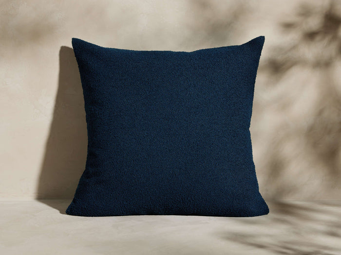 Harborside Outdoor Pillow. Textured navy blue bouclv© square pillow, upright, on a light surface with plant shadows.