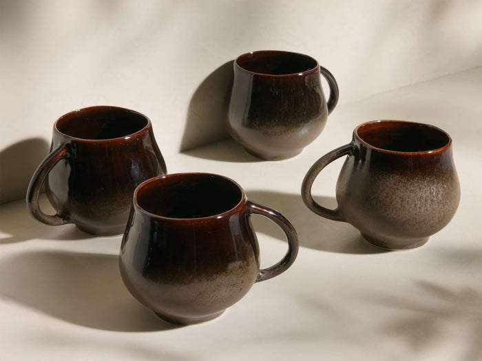 Four Heartline Mugs in dark brown to speckled tan gradient, with rounded bodies and handles, arranged on a light surface.