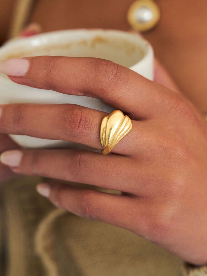 A close-up of a hand wearing a textured gold ring, holding a white ceramic mug against a warm brown background.