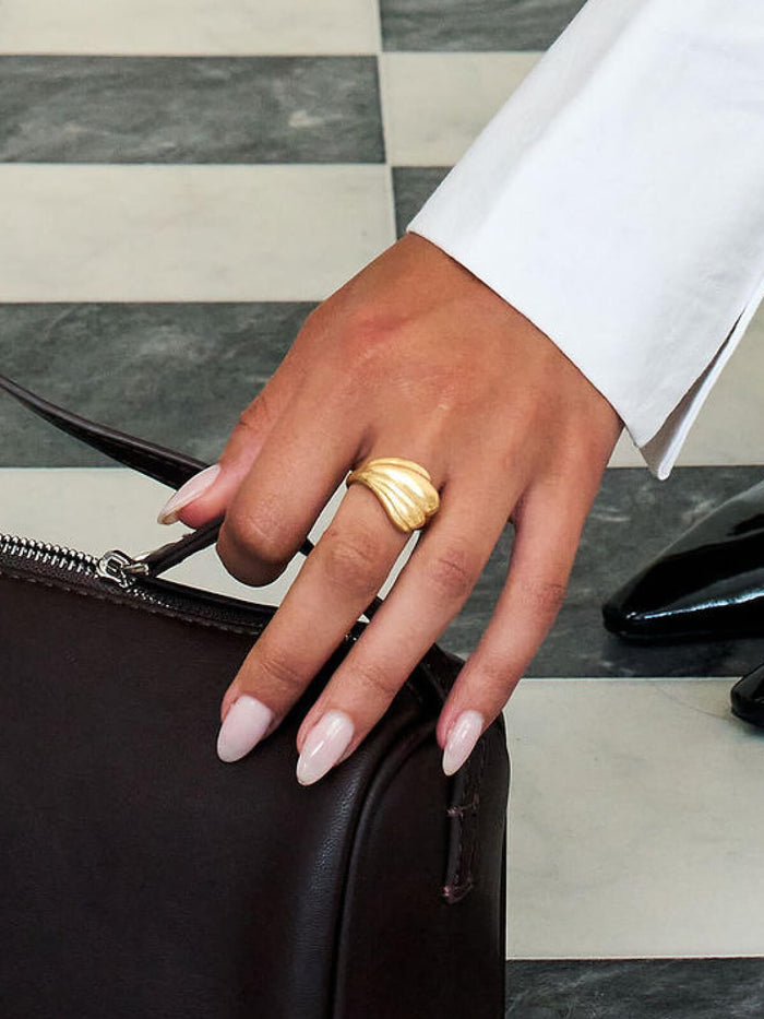 A close-up of a hand wearing a textured gold ring, resting on a dark brown leather bag against a black and white checkered floor.
