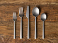 Holmes Five Piece Place Setting: Matte stainless steel forks, knife, and spoons arranged vertically on a rustic wooden table.