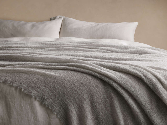 Full view of a light grey Italian Alpaca Boucle Throw, elegantly draped over white bed sheets, showcasing its plush boucle texture.