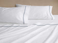 White Italian Embroidered Percale Sheet Set: two pillowcases and flat sheet with navy blue embroidered stripes, casually draped on a bed.