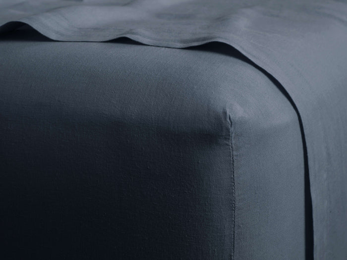 Deep blue-grey Italian Garment-Dyed Linen Fitted Sheet, showing a fitted corner on a mattress with a draped sheet.