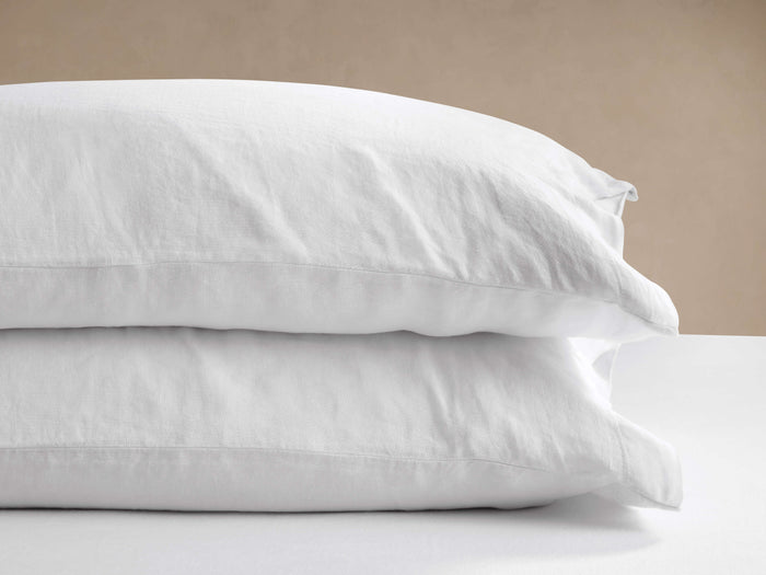 Two white Italian Garment-Dyed Linen Pillowcases, stacked on a white surface with a light beige wall behind them.