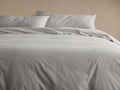 Pale grey Italian Garment-Dyed Percale Duvet, lightly wrinkled, with two matching pillowcases on a bed. Warm wall background.
