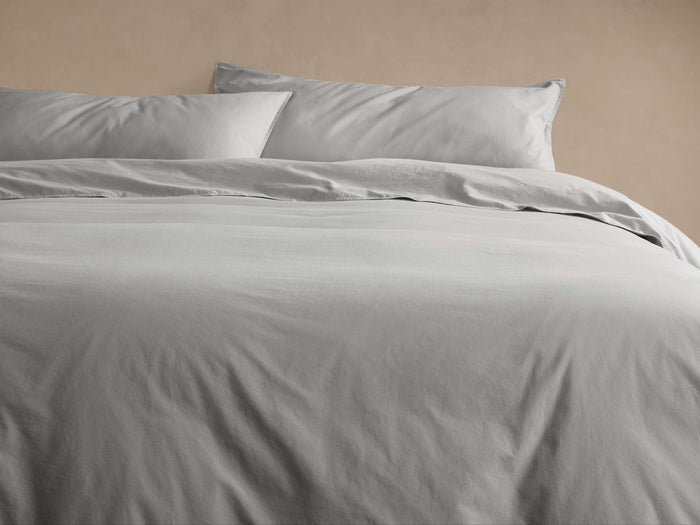 Pale grey Italian Garment-Dyed Percale Duvet, lightly wrinkled, with two matching pillowcases on a bed. Warm wall background.