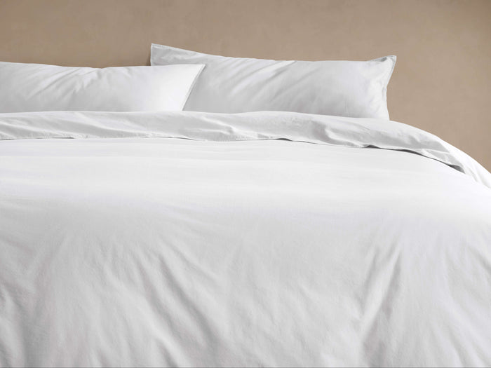 White Italian Garment-Dyed Percale Duvet Set, featuring a relaxed duvet cover and two pillow shams on a bed against a beige wall.
