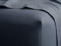 Slate blue Italian Garment-Dyed Percale Fitted Sheet, stretched on a mattress corner, with a flat sheet folded above.