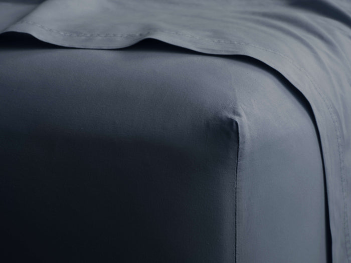 Slate blue Italian Garment-Dyed Percale Fitted Sheet, stretched on a mattress corner, with a flat sheet folded above.