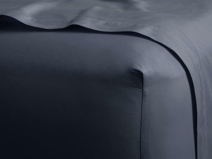 Dark blue Italian Garment-Dyed Sateen Fitted Sheet smoothly covers a bed corner, a sheet drapes over it.