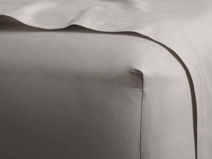 Light grey Italian Garment-Dyed Sateen Fitted Sheet on a bed corner, showing smooth fabric & subtle sheen. Another sheet drapes over.