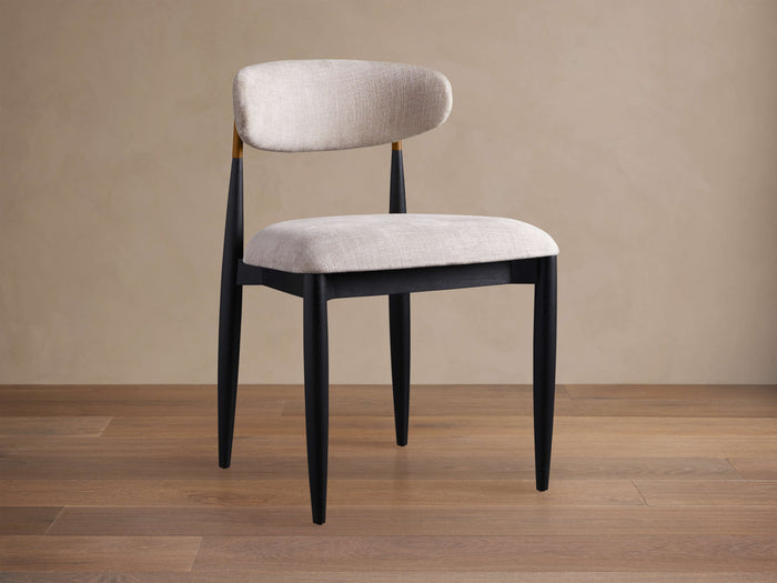 Jagger Dining Side Chair, angled, with black frame, light beige upholstered seat & curved back, gold accent, on wood floor.
