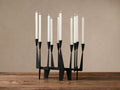 Matte black iron Javari Candleholder with unique geometrical silhouette, holding 8 white taper candles, some lit, on a rustic wood table.