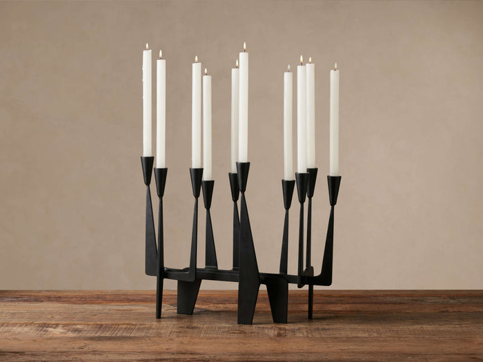 Matte black iron Javari Candleholder with unique geometrical silhouette, holding 8 white taper candles, some lit, on a rustic wood table.