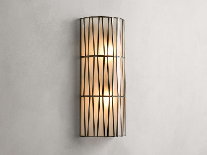 Jeunet Sconce with antiqued brass finish, features two glowing lights behind a cylindrical iron frame with vertical and diagonal lines.