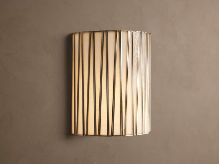Jeunet Sconce, glowing, with vertical antiqued brass iron strips over translucent white glass, mounted on a neutral wall.
