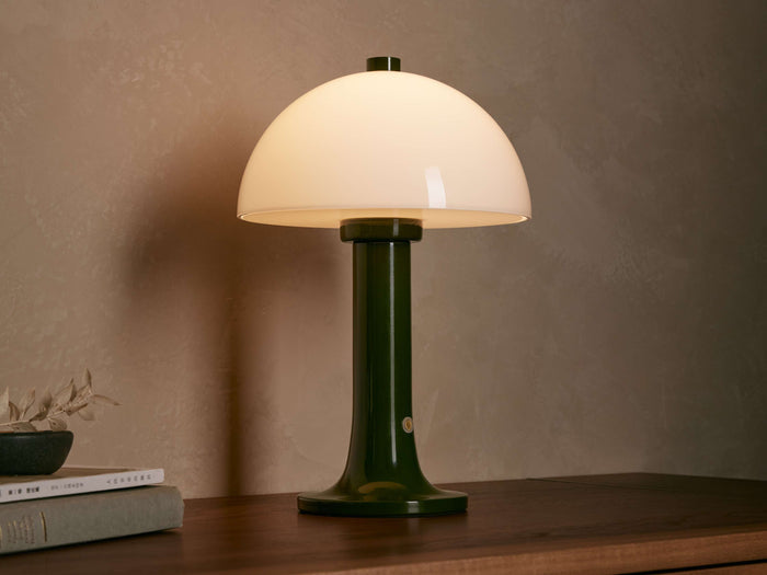 Jocy Portable Table Lamp with a dark green lacquered base and a domed white opal shade, illuminated on a wooden table.