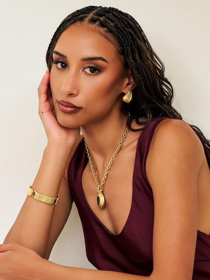 A woman with braided hair wearing a gold chain necklace with a teardrop pendant, a gold bangle, and a gold hoop earring, paired with a dark plum sleeveless top against a plain light background.