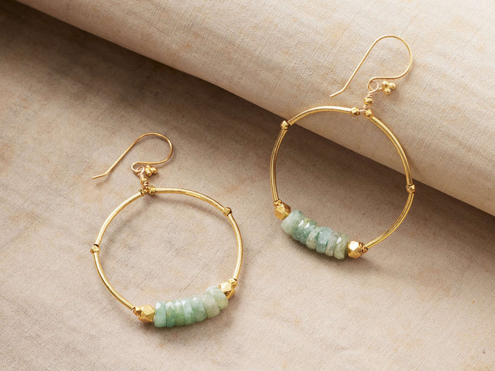 Kelby Hoop Earrings: Two gold vermeil hoops with pale green faceted aquamarine stones and French-wire backs on a textured surface.