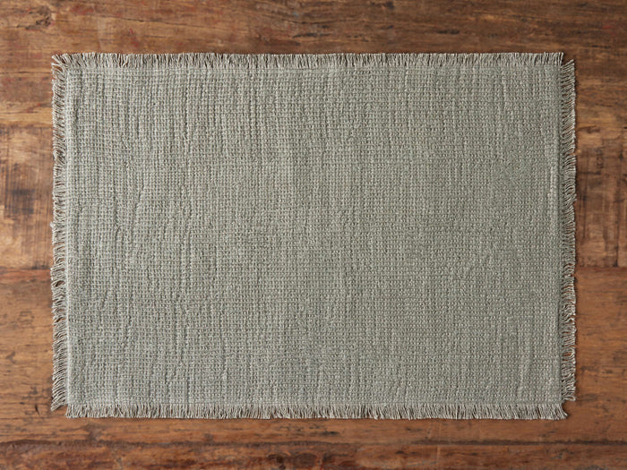 Khai Placemat shown on a wooden surface, featuring a woven texture in warm neutral tones with frayed edges for a rustic look.