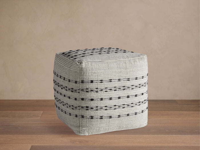 Ladera Performance Pouf with a square shape, featuring a grey and black striped pattern, on a wooden floor against a neutral wall.