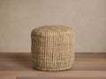 Laos Jute Pouf. Light brown, cylindrical, woven jute pouf on a wooden floor against a light beige wall.