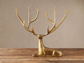 Laying Deer Statue with antiqued brass finish, large branched antlers, posed facing forward on a rustic wooden surface.