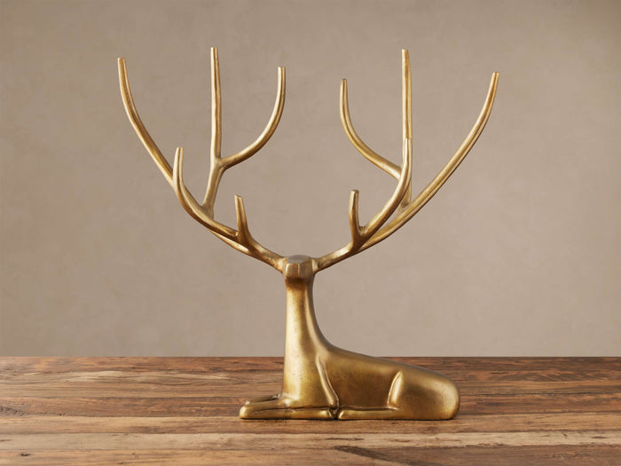 Laying Deer Statue with antiqued brass finish, large branched antlers, posed facing forward on a rustic wooden surface.