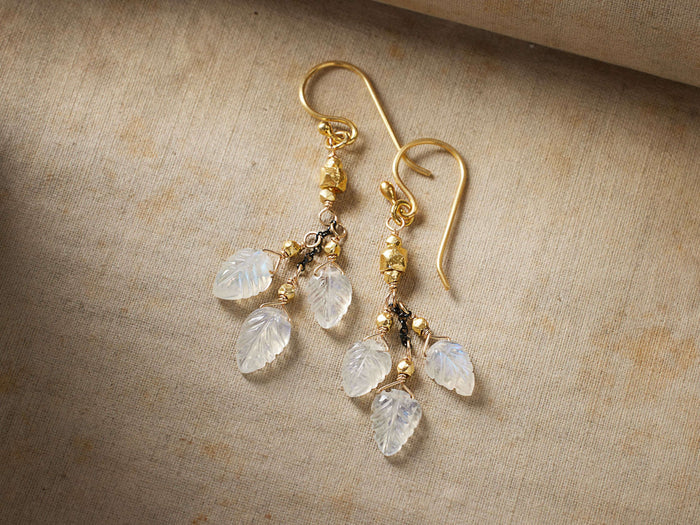 Leaf Drop Earrings with carved moonstone leaves hanging from 14 kt gold ear wires, adorned with gold beads, on a textured surface.