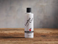 White "Leather Care Conditioner" bottle with black cap & red apple logo, standing upright on a rustic wooden surface with a grey background.