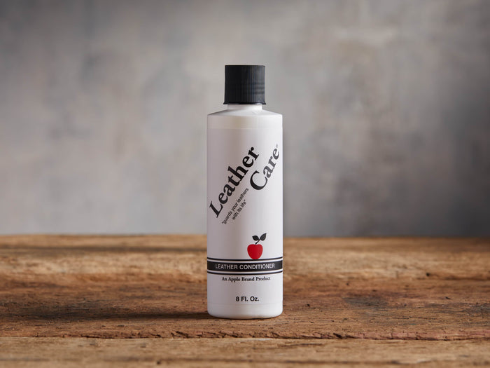 White "Leather Care Conditioner" bottle with black cap & red apple logo, standing upright on a rustic wooden surface with a grey background.