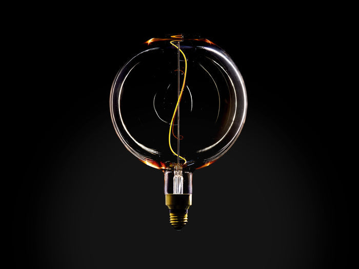 Clear glass globe bulb with amber tint and visible yellow filament inside. E26 base at bottom. Dark background highlighting bulb's design.