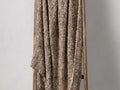 Leni Throw in a textured brown and beige wool blend, draped over a wooden ladder. Fringed edges visible on one side.