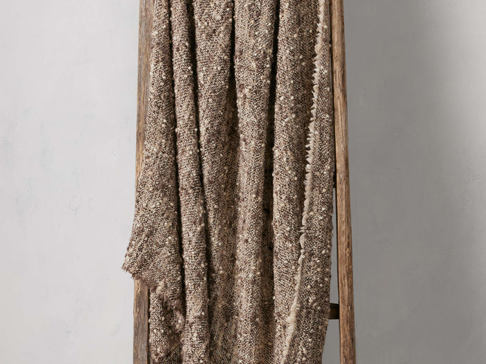Leni Throw in a textured brown and beige wool blend, draped over a wooden ladder. Fringed edges visible on one side.