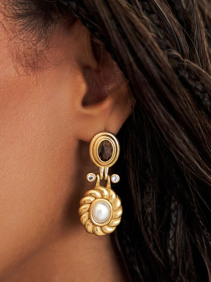 A close-up of a model's ear wearing a gold drop earring with a central white pearl and a small dark oval stone above it.